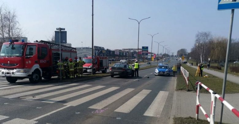 Zdjęcie z miejsca wypadku, na pierwszym planie oznakowane przejście dla pieszych, w tle radiowóz, policjanci, wóz strażacki i strażacy