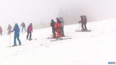 Zamglone, ośnieżone zbocze góry. W grupie stoi sześciu narciarzy w kolorowych kombinezonach