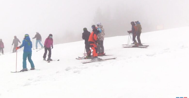 Zamglone, ośnieżone zbocze góry. W grupie stoi sześciu narciarzy w kolorowych kombinezonach