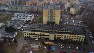 Na zdjęciu budynek widoczny z lotu ptaka. Żółta kondygnacja, szary dach, wokół budynku parkingi z zaparkowanymi samochodami