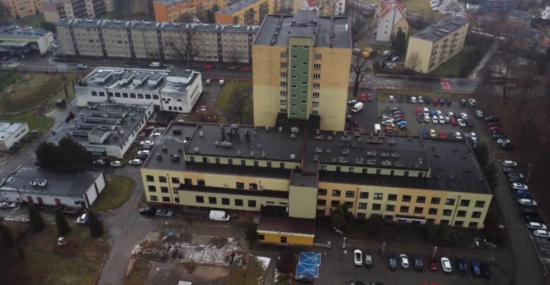 Na zdjęciu budynek widoczny z lotu ptaka. Żółta kondygnacja, szary dach, wokół budynku parkingi z zaparkowanymi samochodami