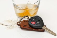 Na zdjęciu stłuczona szklanka z żółtym napojem alkoholowym, obok leżą kluczyki do samochodu z przyciskami do centralnego zamka, dopięte do skórzanego, prostokątnego breloka