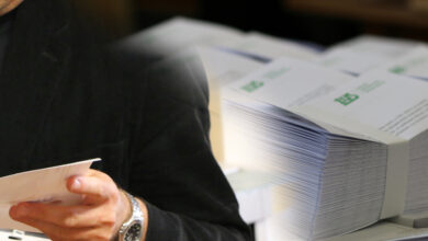 Dodatkowa emerytura - grafika, fragment postaci, ręce trzymające dokument w tle stosy kopert z listami do doręczenia