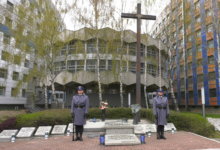 Grób Policjanta Polskiego. Dwaj Policjanci w niebieskich mundurach z bronią przy nodze trzymająstrażprzy grobie. W tle dwa drzewa i budynki