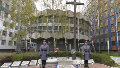 Grób Policjanta Polskiego. Dwaj Policjanci w niebieskich mundurach z bronią przy nodze trzymająstrażprzy grobie. W tle dwa drzewa i budynki