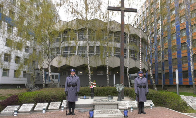 Grób Policjanta Polskiego. Dwaj Policjanci w niebieskich mundurach z bronią przy nodze trzymająstrażprzy grobie. W tle dwa drzewa i budynki