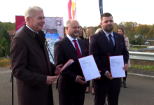 Trasa N-s Trzech mężczyzn trzyma w rękach papierowe teczki pozuja do zdjęcia w tle kolorowe flagi i stojący ludzie