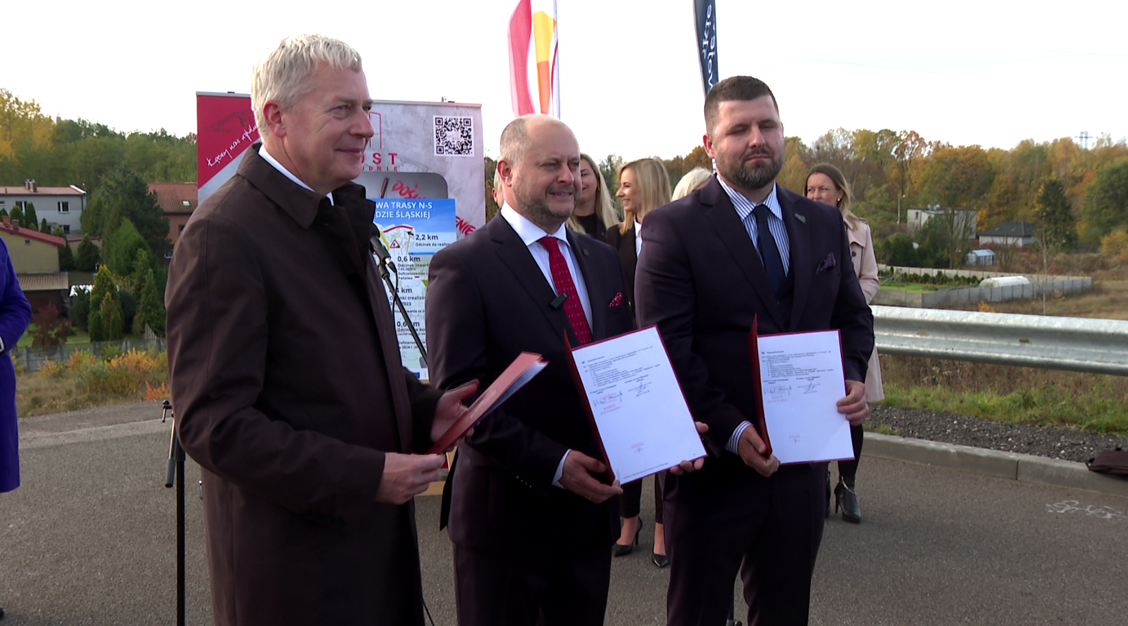 Trasa N-s Trzech mężczyzn trzyma w rękach papierowe teczki pozuja do zdjęcia w tle kolorowe flagi i stojący ludzie